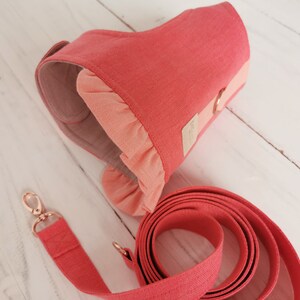 May include: A coral pink dog harness with a ruffled trim and a matching leash. The harness has a gold metal ring for attaching a leash.