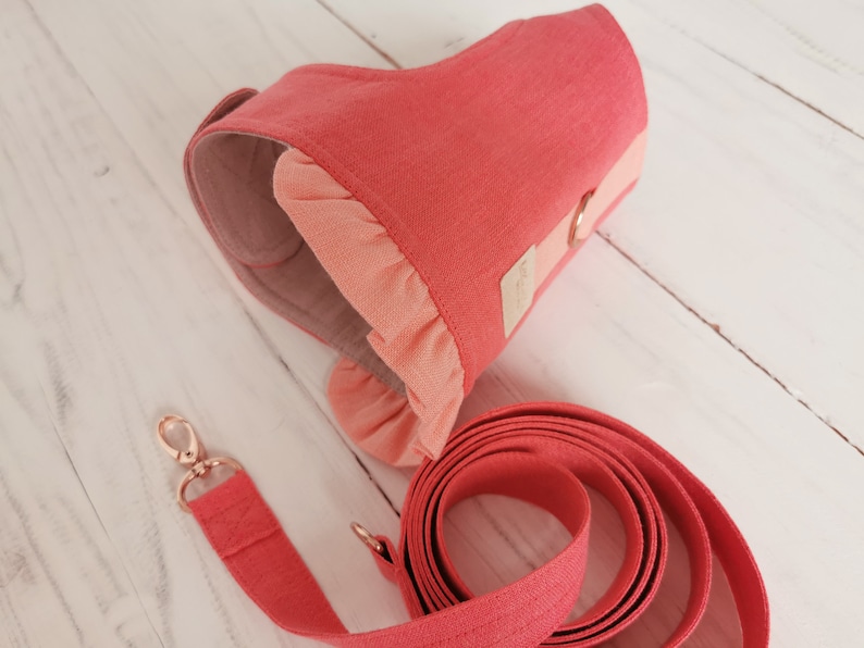 May include: A coral pink dog harness with a ruffled trim and a matching leash. The harness has a gold metal ring for attaching a leash.
