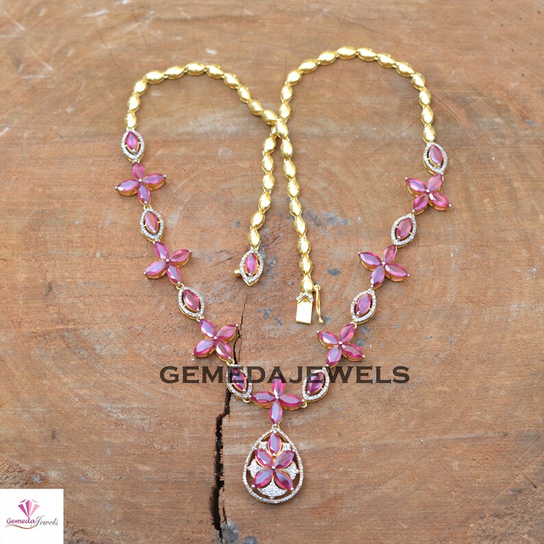 May include: A gold necklace with a pink gemstone pendant and pink gemstone flowers. The necklace is on a wooden surface. GEMEDAJEWELS is printed on the surface.