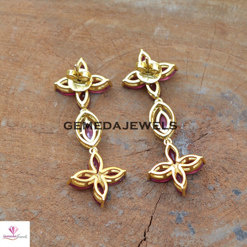 May include: A pair of gold earrings with pink gemstones. The earrings are shaped like a flower with four petals. The earrings are dangling and have a small loop at the top for attaching to an earring post.