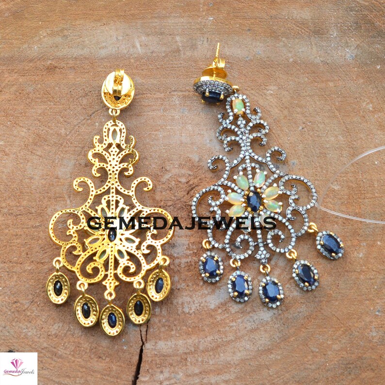 May include: A pair of gold and silver dangle earrings with a floral design. The earrings feature blue and white gemstones and the text "GEMEDAJEWELS".
