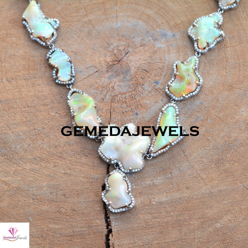 May include: A silver necklace with a large, irregular-shaped opal pendant and smaller opal stones. The necklace is set with small diamonds and has a delicate, feminine design. GEMEDAJEWELS is printed on the image.