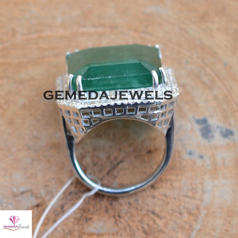 May include: A silver ring with a large, emerald green gemstone set in a square, open-work design. The ring is surrounded by smaller, white gemstones.