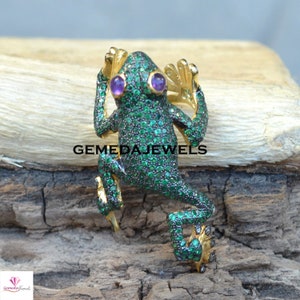 Puede incluir: Una figura de rana enjoyada con gemas verdes que cubren su cuerpo y detalles dorados en las patas y manos. La rana tiene ojos morados y está posando como si estuviera trepando. El texto "GEMEDA JEWELS" está impreso en la imagen.