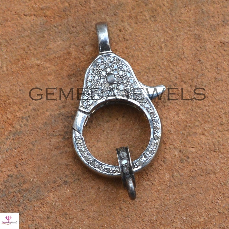 May include: A silver clasp with a round opening and a hook closure. The clasp is covered in small, clear diamonds.