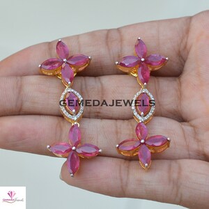 May include: A pair of dangle earrings with a floral design. The earrings are made of gold and feature red ruby stones and clear diamonds. The earrings are approximately 2 inches long.