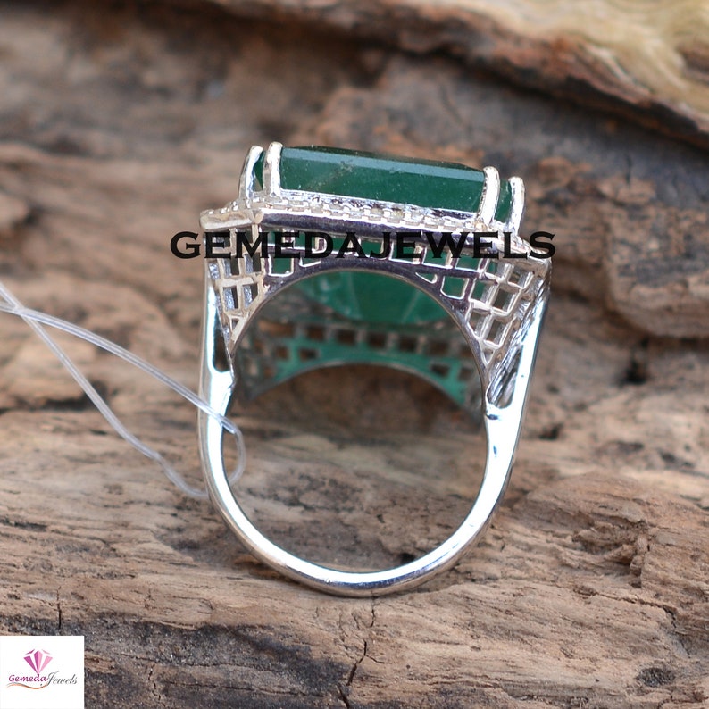 May include: A silver ring with a large, emerald green gemstone set in a cage-like design. The ring is on a wooden surface. GEMEDAJEWELS is printed on the ring.