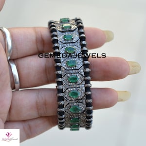 May include: A silver bracelet with emeralds and small diamonds. The bracelet has a black cord design between the silver sections. The bracelet has the text "GEMEDA JEWELS" on it.