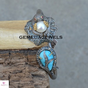 Könnte beinhalten: Ein silbernes Armband mit einem türkisfarbenen Stein und einer Perle, die von einem silbernen und schwarzen Design umgeben ist. Das Armband ist mit kleinen Diamanten verziert. Ein am Armband befestigtes Etikett trägt die Aufschrift GEMEDA JEWELS.