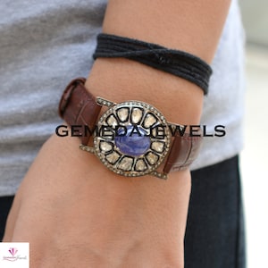 May include: A silver watch with a blue gemstone surrounded by diamonds. The watch has a brown leather band and the text "GEMEDAL JEWELS" is visible on the band.