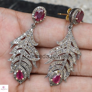 May include: A pair of silver feather earrings with a red ruby gemstone at the bottom of each earring. The earrings are adorned with small diamonds.