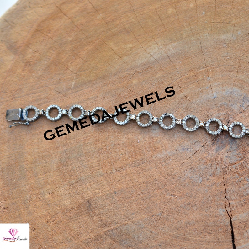 May include: A silver bracelet with a circle design, each circle is set with small diamonds. The bracelet is on a wooden surface. GEMEDA JEWELS is written on the surface.