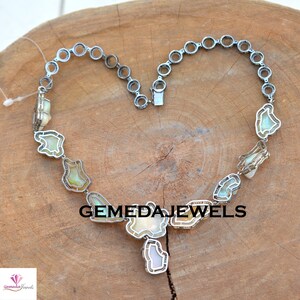 May include: A silver necklace with a heart-shaped design featuring a large iridescent gemstone pendant. The necklace is made of silver and has a chain with small round links. The gemstone is surrounded by smaller gemstones. The necklace is on a wooden surface. GEMEDAJEWELS is printed on the surface.