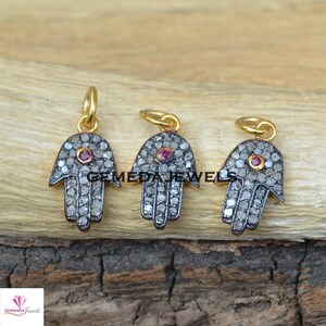May include: Three silver hamsa hand charms with pave diamond accents and a single red gemstone in the center of each charm. The charms are on a wooden surface.