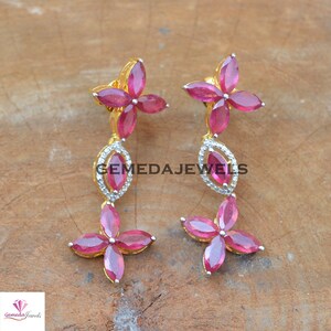 May include: A pair of dangle earrings with a floral design. The earrings are made of gold and feature pink gemstones and clear cubic zirconia stones. The earrings are accented with a small, clear cubic zirconia stone in the center of each flower.