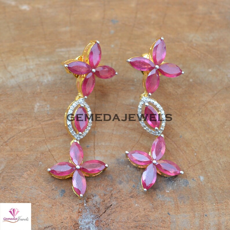 May include: A pair of dangle earrings with a floral design. The earrings are made of gold and feature pink gemstones and clear cubic zirconia stones. The earrings are accented with a small, clear cubic zirconia stone in the center of each flower.