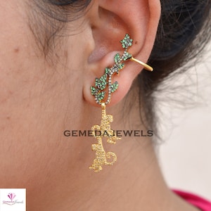 May include: Gold ear cuff with a leaf design featuring green and black gemstones. The ear cuff is attached to a dangling earring with a similar leaf design.