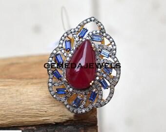 Könnte beinhalten: Ein silberner Ring mit einem roten Edelstein und blauen Baguette-förmigen Edelsteinen, die in einem floralen Design eingefasst sind.