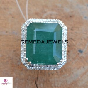 May include: A silver ring with a large emerald green gemstone surrounded by a halo of smaller diamonds. The ring is set in a square shape with a beveled edge. The text "GEMEDAJEWELS" is visible on the ring.
