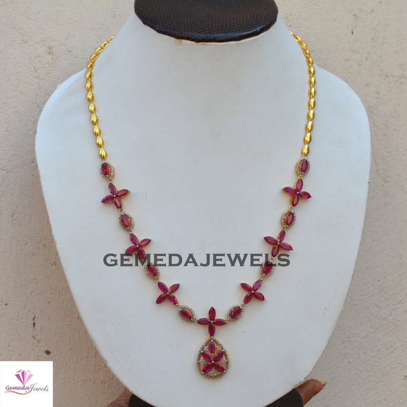 May include: A gold necklace with a pendant featuring a red ruby flower design. The necklace is made of small gold beads and features red ruby flowers with white diamond accents. The pendant is a teardrop shape with a red ruby flower in the center.