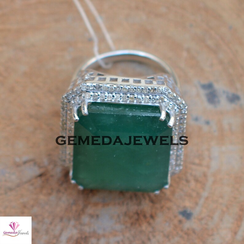 May include: A silver ring with a large, emerald green gemstone surrounded by smaller, clear gemstones. The ring is set in a square-shaped setting with a beveled edge. The text "GEMEDAJEWELS" is visible on the ring.