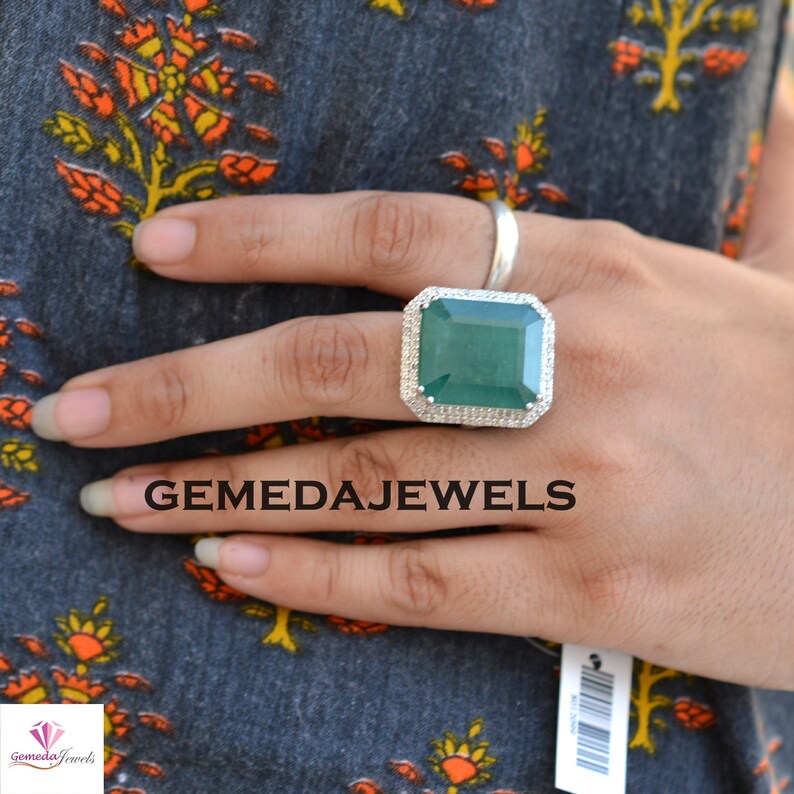 May include: A silver ring with a large, square, emerald green gemstone surrounded by smaller, clear gemstones. The ring is on a person's finger.