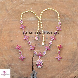 May include: A gold necklace and earrings set with a pink gemstone pendant and matching earrings. The necklace features a chain with pink gemstone flowers and a teardrop-shaped pendant with a white gemstone center. The earrings are dangle earrings with pink gemstone flowers.