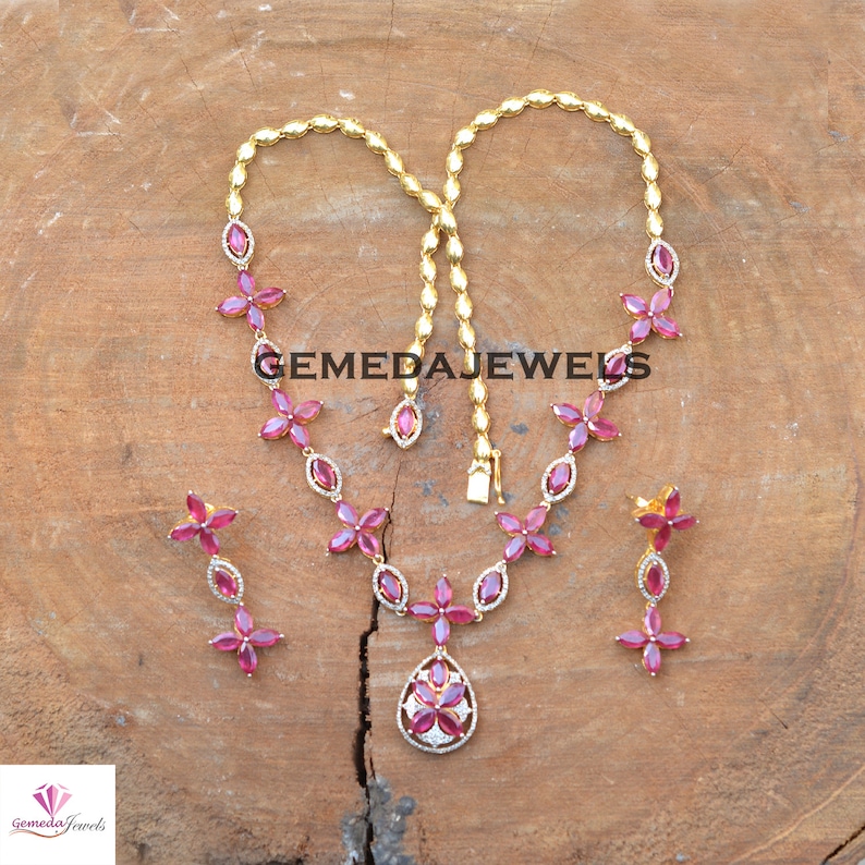May include: A gold necklace and earrings set with a pink gemstone pendant and matching earrings. The necklace features a chain with pink gemstone flowers and a teardrop-shaped pendant with a white gemstone center. The earrings are dangle earrings with pink gemstone flowers.