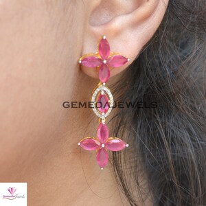 May include: Gold dangle earrings with pink gemstones and clear cubic zirconia stones. The earrings feature a flower design with five petals.