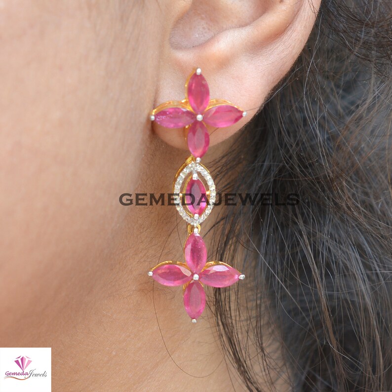 May include: Gold dangle earrings with pink gemstones and clear cubic zirconia stones. The earrings feature a flower design with five petals.