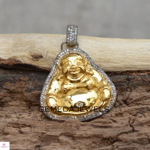 Puede incluir: Un colgante de color dorado con una figura de Buda sonriente, rodeado por un borde de pequeños diamantes transparentes. El colgante está hecho de plata y tiene un aro en la parte superior para sujetarlo a una cadena.