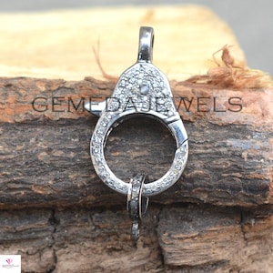 May include: Silver clasp with a round opening and a small loop at the bottom. The clasp is covered in small, clear diamonds.