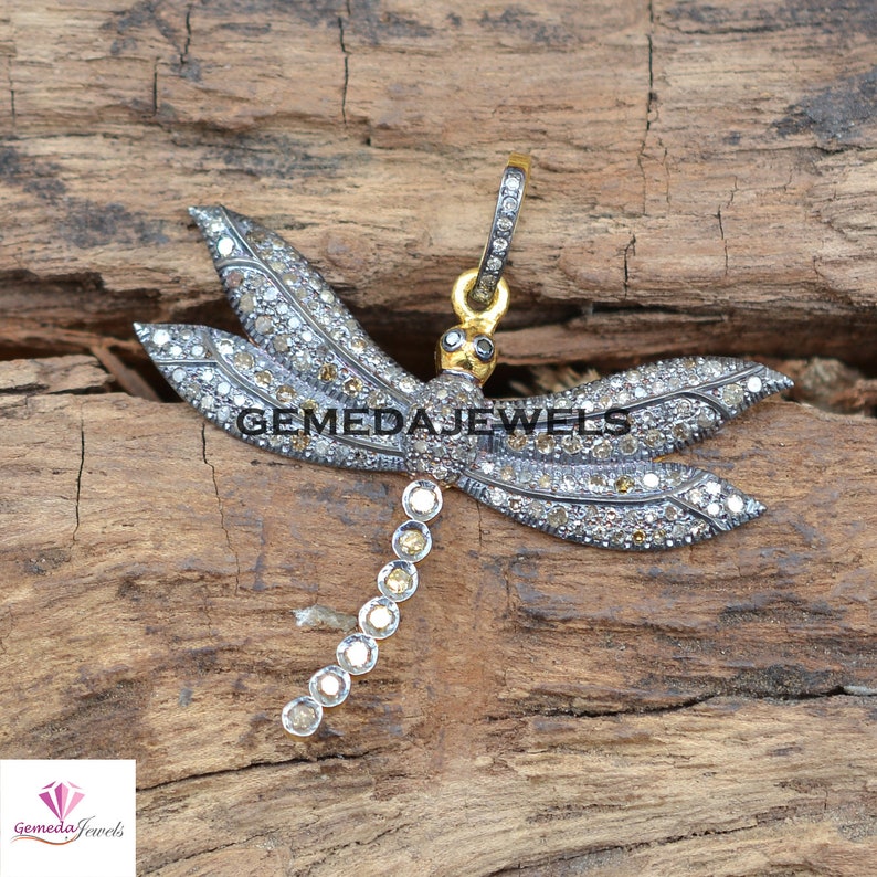 Peut inclure: Un pendentif en argent en forme de libellule avec une cha&icirc;ne en or et des diamants clairs et bruns. Le pendentif est plac&eacute; sur un fond en bois.