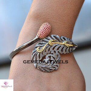 Op de afbeelding: Een zilverkleurige manchetarmband met een veermotief. De veer is versierd met heldere edelstenen en gouden accenten. De armband heeft een roze edelsteengroep en de tekst "GEMEDA JEWELS".