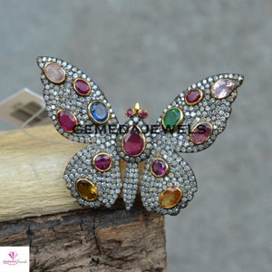 Op de afbeelding: Een zilveren vlinder broche met een pavé setting van kleine diamanten en gekleurde edelstenen, waaronder rood, roze, groen, blauw en geel.
