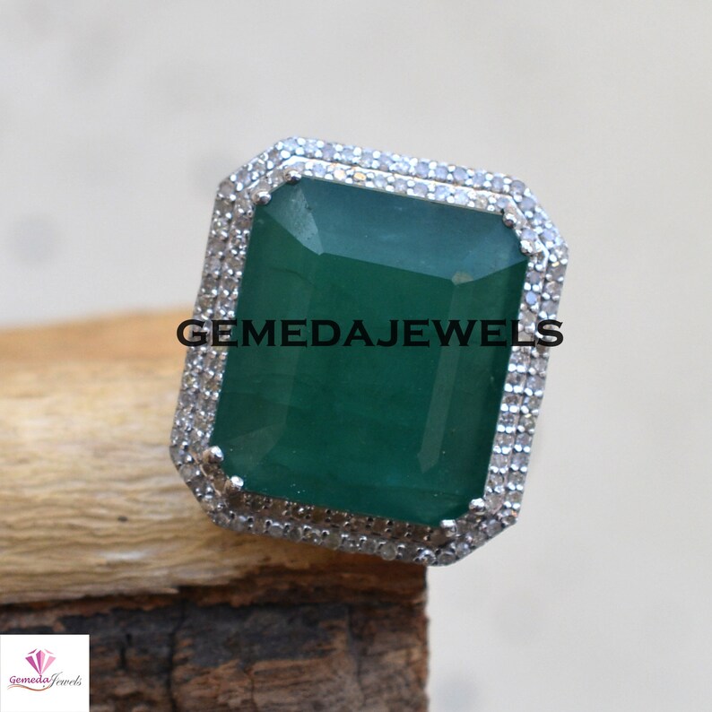 May include: A silver ring with a large, emerald green gemstone surrounded by a double row of smaller, clear gemstones. The ring is set in a square-shaped setting. The text "GEMEDAJEWELS" is visible on the ring.