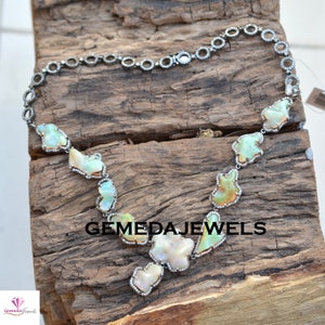 May include: A silver necklace with a chain made of small circles and a pendant made of several irregular shaped opal stones. The opal stones are set in silver and have a white, green, and orange color. The necklace is on a wooden surface. GEMEDAJEWELS is written in black text on the surface.