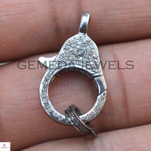 May include: A silver clasp with a round ring and a smaller ring attached. The clasp is covered in small, white diamonds.