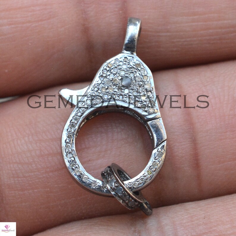 May include: A silver clasp with a round ring and a smaller ring attached. The clasp is covered in small, white diamonds.