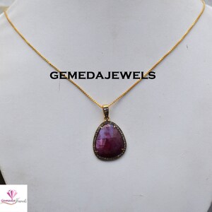 Pode incluir: Um colar de corrente de ouro com um pingente que apresenta uma grande gema oval de cor rosa escura cercada por um halo de gemas brancas menores. O pingente é engastado em uma moldura de ouro. O texto "GEMEDAJEWELS" é visível acima do pingente.
