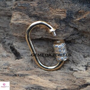 Puede incluir: Cierre de metal dorado con un dije cil&iacute;ndrico incrustado con peque&ntilde;as piedras transparentes. El dije est&aacute; grabado con el texto "GEMEDAJEWELS".