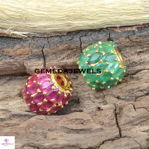 May include: Two round gold beads with a hole through the center. One bead is set with emerald green gemstones and the other is set with ruby red gemstones.