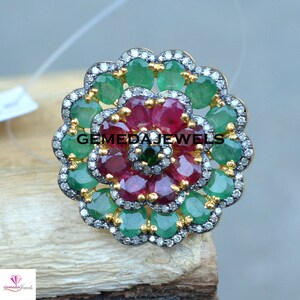 May include: A silver ring with a flower design featuring red rubies and green emeralds surrounded by diamonds. The ring is set on a wooden surface. GEMEDA JEWELS is printed on the ring.