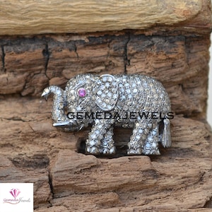 May include: A silver elephant brooch with a pink gemstone eye, covered in small diamonds. The brooch is on a wooden surface.