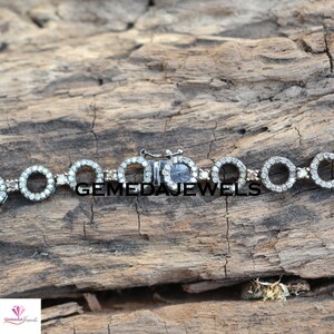 May include: A silver bracelet with a clasp closure. The bracelet is made of small circles that are set with diamonds. The bracelet is on a wooden surface.