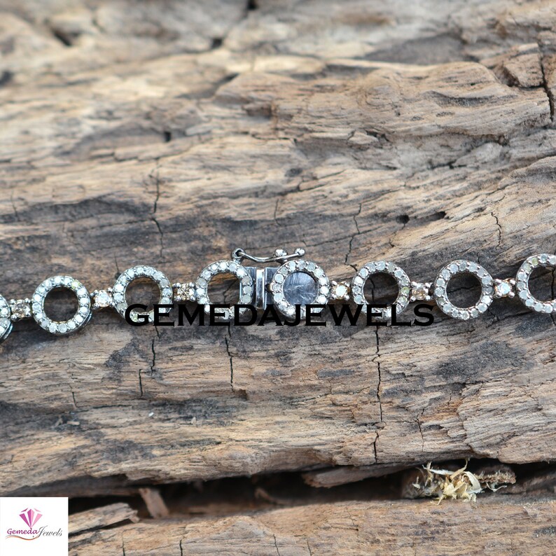 May include: A silver bracelet with a clasp closure. The bracelet is made of small circles that are set with diamonds. The bracelet is on a wooden surface.