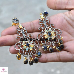 May include: A pair of silver dangle earrings with a floral design. The earrings are adorned with white opals and blue sapphires. The earrings are approximately 3 inches long.