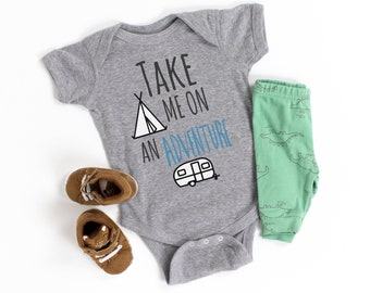 Könnte beinhalten: Ein grauer Baby-Strampler mit dem Text "Take me on an adventure" in Schwarz und Blau. Der Strampler zeigt eine Schwarz-Weiß-Illustration eines Zeltes und eines Wohnwagens. Der Strampler wird mit einer grünen Hose und braunen Schuhen kombiniert.