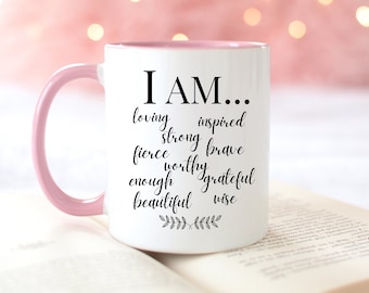 May include: White ceramic mug with a pink rim and handle. The mug has black text that reads "I AM..." followed by a list of positive affirmations: loving, inspired, strong, brave, fierce, worthy, enough, grateful, beautiful, wise. The mug is sitting on a book.