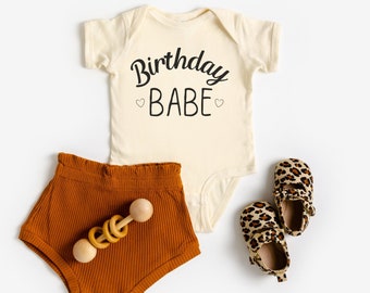 May include: A beige baby onesie with the text "Birthday Babe" in black script. The onesie is paired with brown corduroy shorts and leopard print baby shoes.